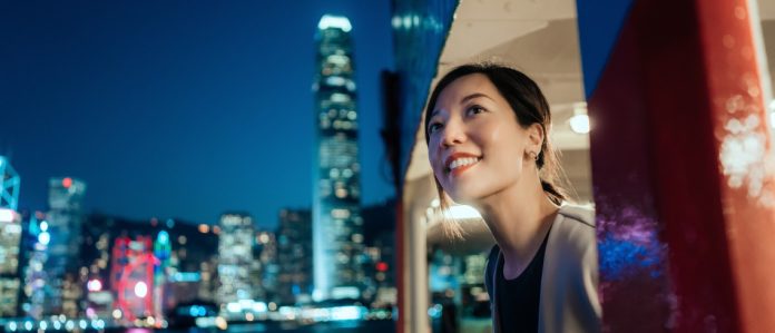 Beautiful smiling young Asian woman looking out through the window, enjoying spectacular illuminated night view of the city while travelling by ferry